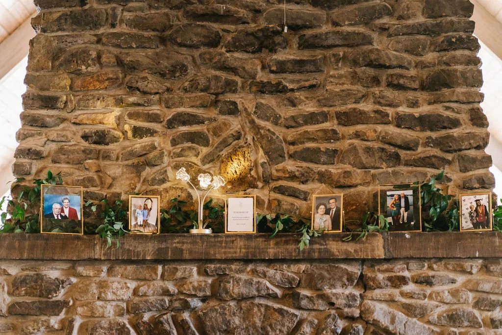 A stone fireplace mantel decorated with framed family photos, a small white flower arrangement, and greenery.