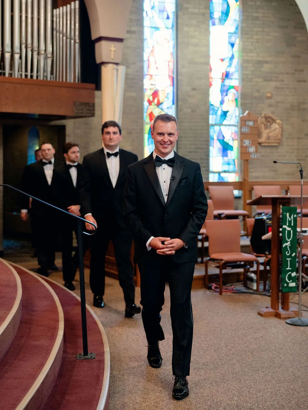 A group of men dressed in formal tuxedos walking inside a church, with stained glass windows in the background and a 'PRESIDENT' sign near the front.