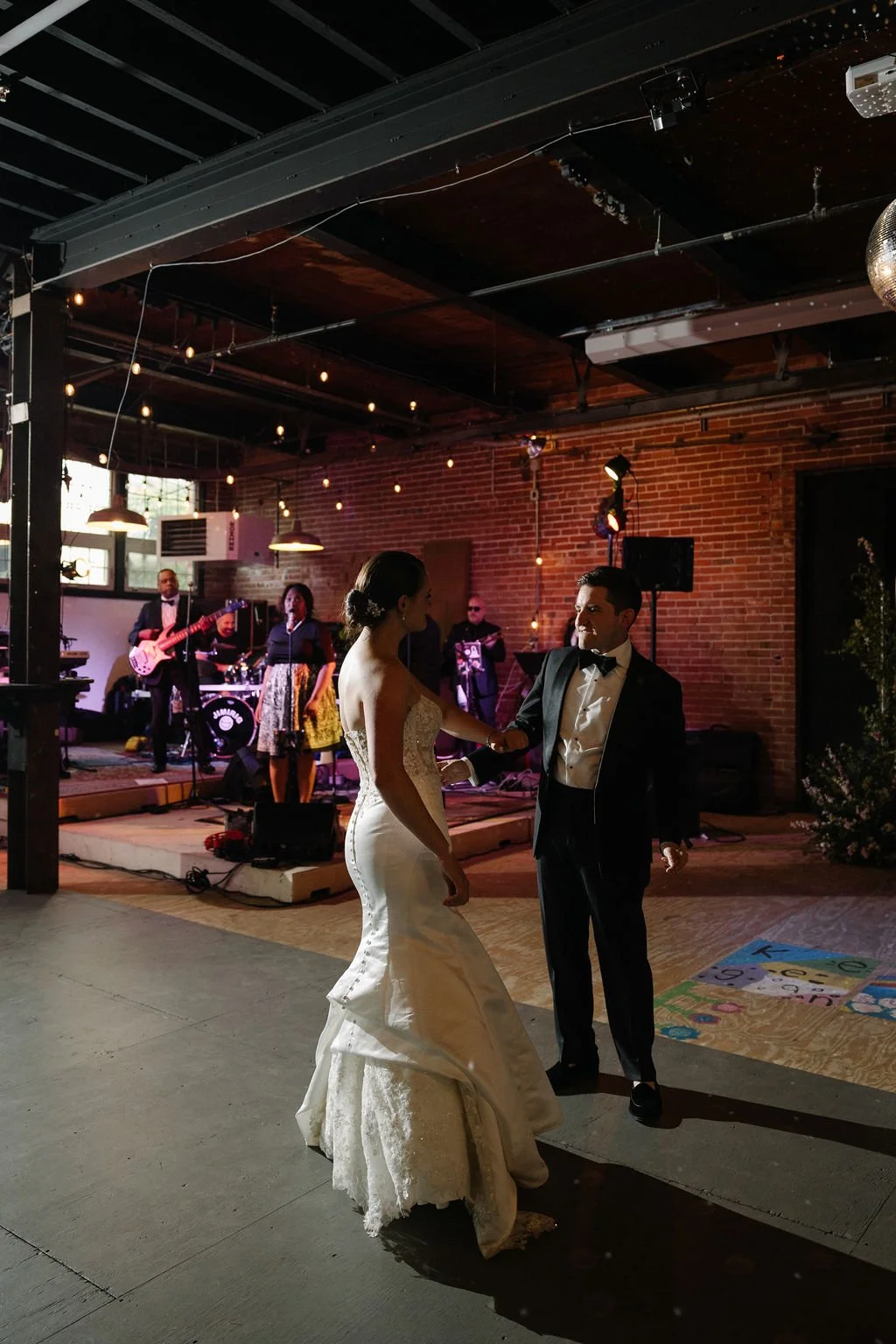 A bride and groom dancing at their wedding reception with a live band playing in the background inside a brick-walled venue.