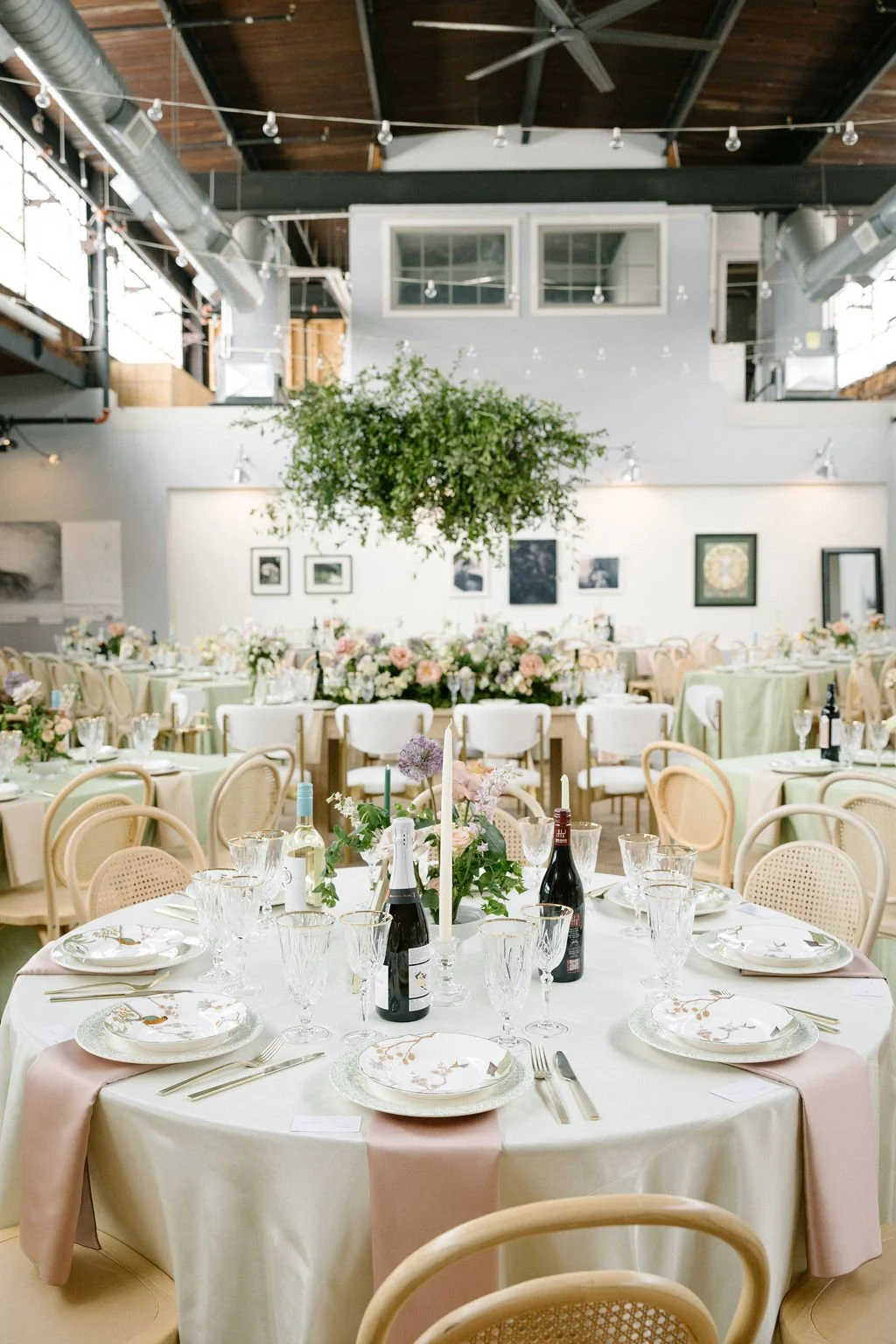 Elegant indoor event space decorated with round tables covered in cream and pastel pink tablecloths, floral centerpieces, wine bottles, glassware, and china, with a large hanging greenery decoration and framed artwork on the walls.