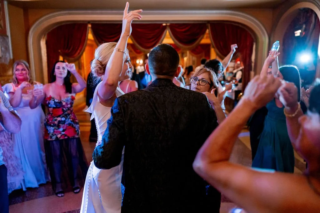 People dancing and celebrating at a formal event in an elegant ballroom with red velvet curtains and warm lighting.