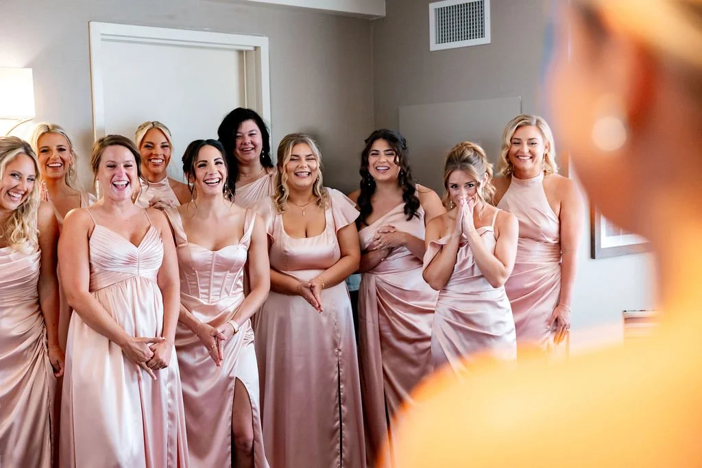 A group of women in light pink dresses smiling and laughing together, likely at a wedding or special event.
