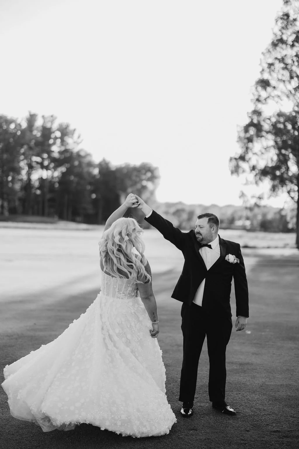 Black and white photo of a couple dancing outdoors, with the woman wearing a wedding dress and the man in a tuxedo, holding hands and dancing in a field with trees in the background.