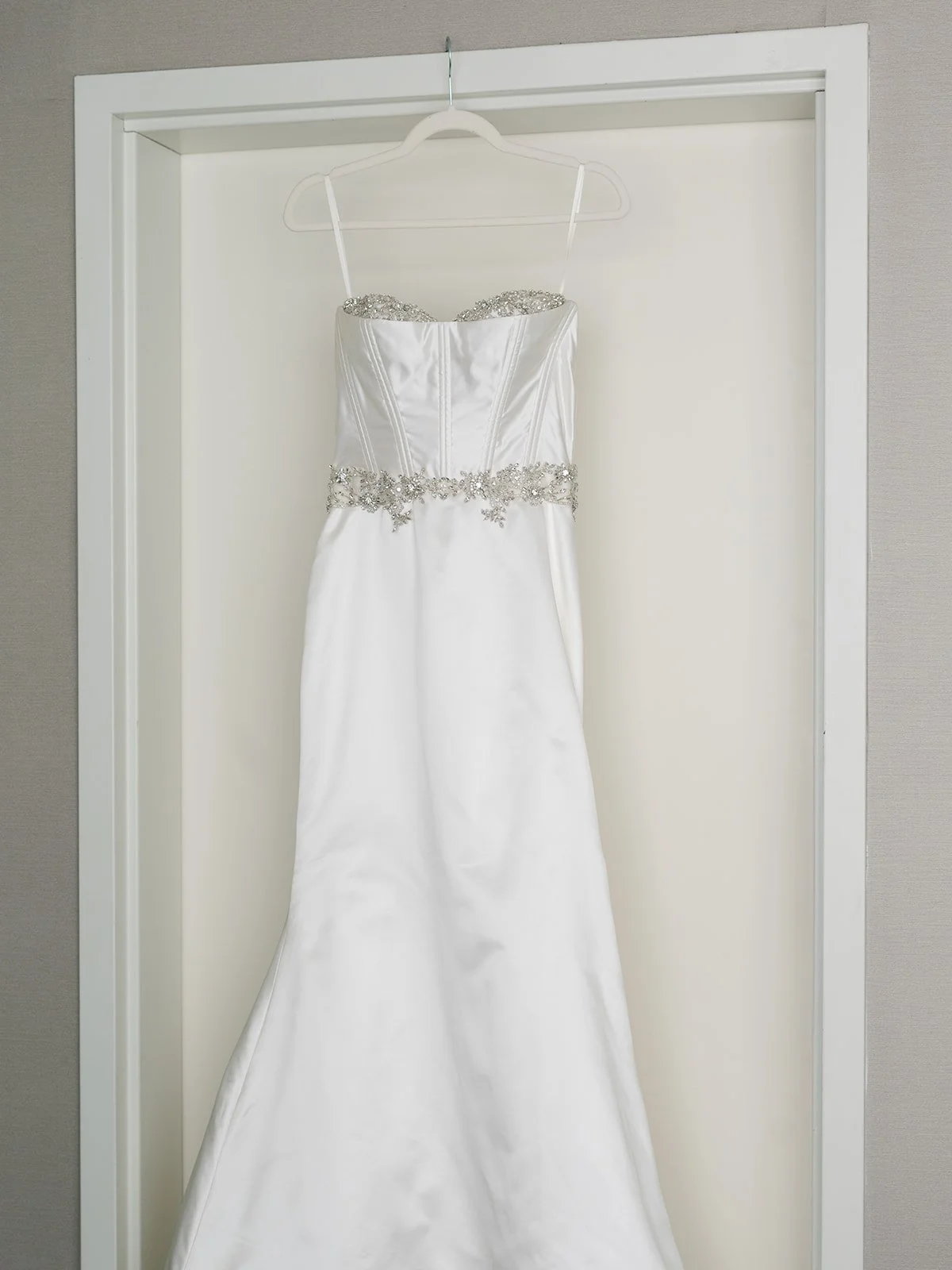 Wedding dress hanging on a white hanger on a door frame, made of satin with beaded embellishments at the waist and neckline.
