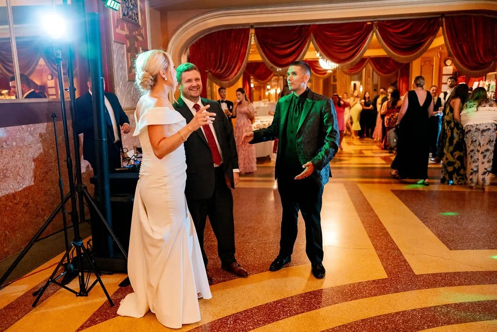Three people dancing and talking at a wedding reception with red drapes and other guests in the background.