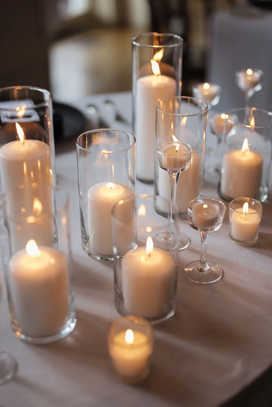 A table decorated with multiple lit white candles in various glass holders, creating a warm and elegant ambiance.