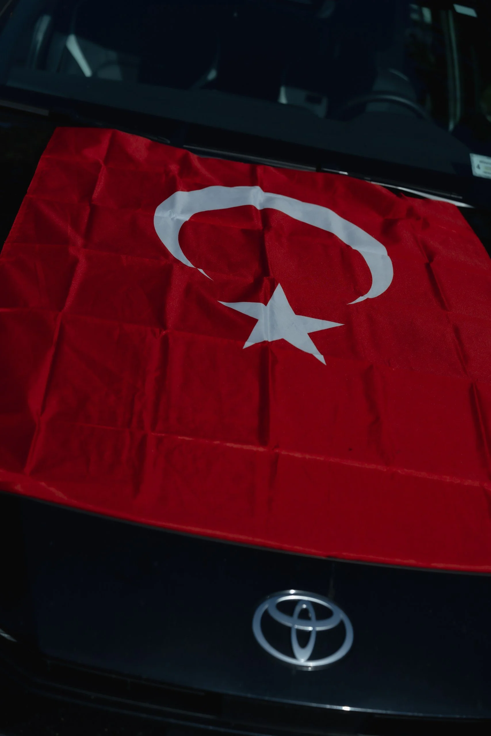 A red flag with a white crescent moon and star draped over the hood of a black Toyota car.
