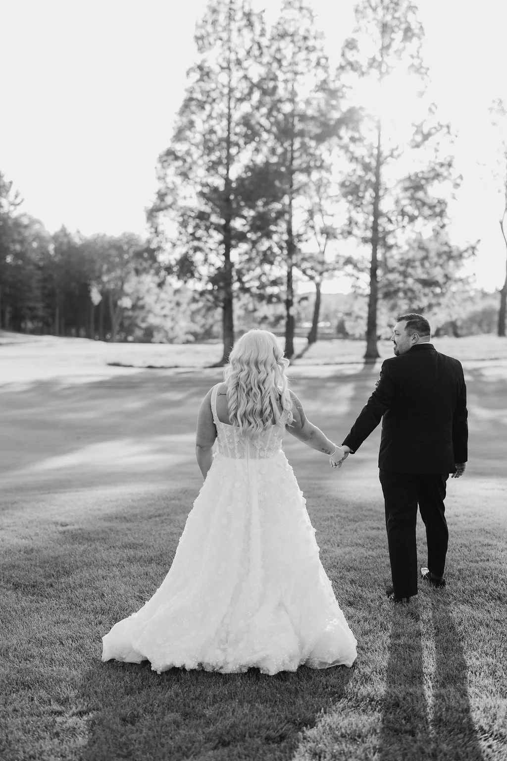 A black-and-white photo of a newlywed couple walking hand-in-hand across a grassy area on a sunny day, with trees in the background.