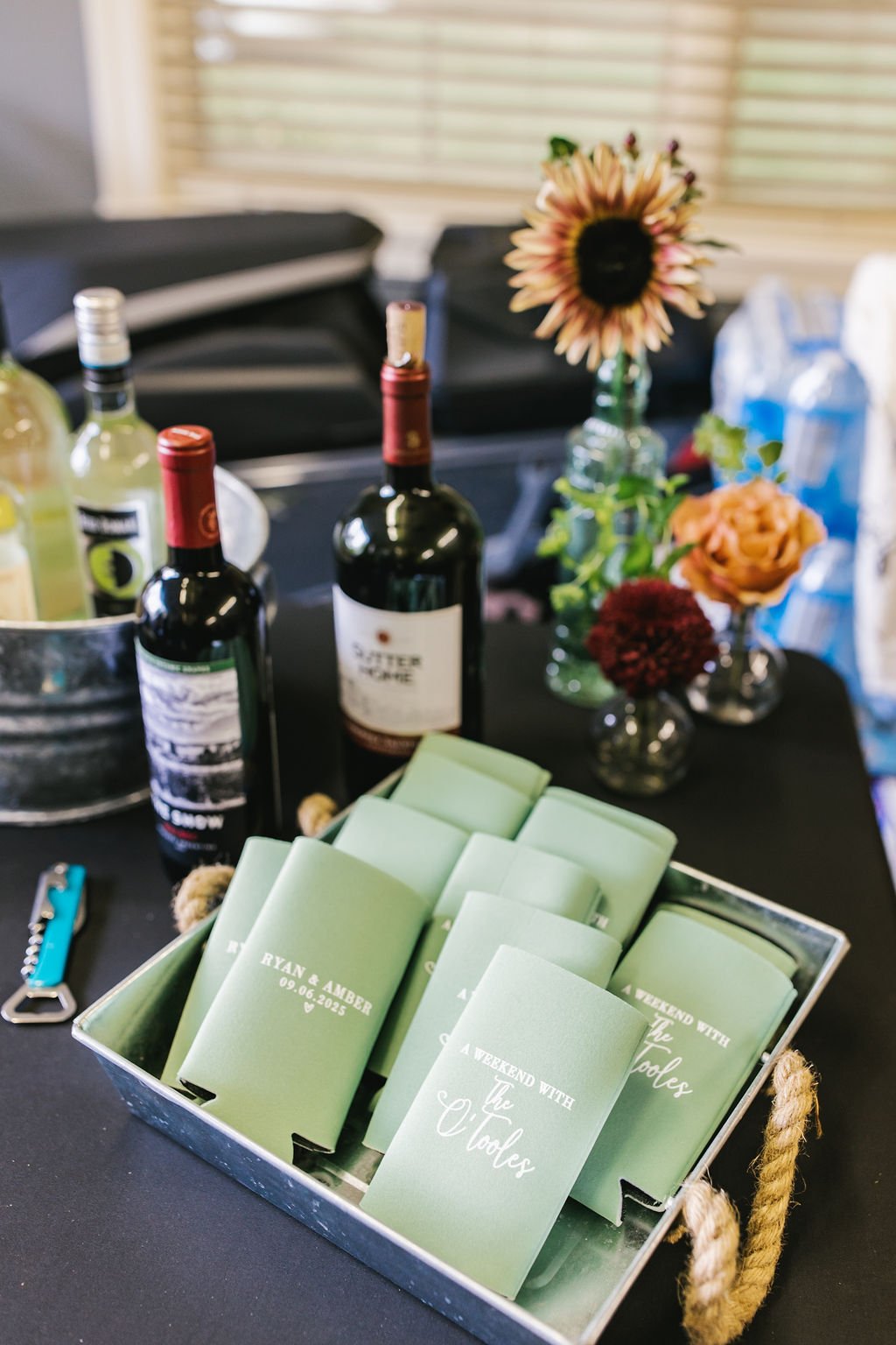 A table with bottles of wine, a small bucket with drinks, flowers in vases, and a box of personalized napkins reading 'Ryan & Amber 09.06.2023 A Weekend with the Fools'.