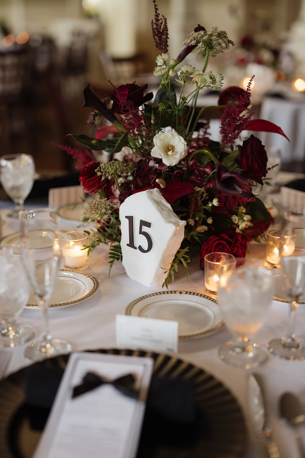 Elegant table centerpiece with dark red, white, and green flowers, surrounded by lit candles and set tableware, with a table number 15.