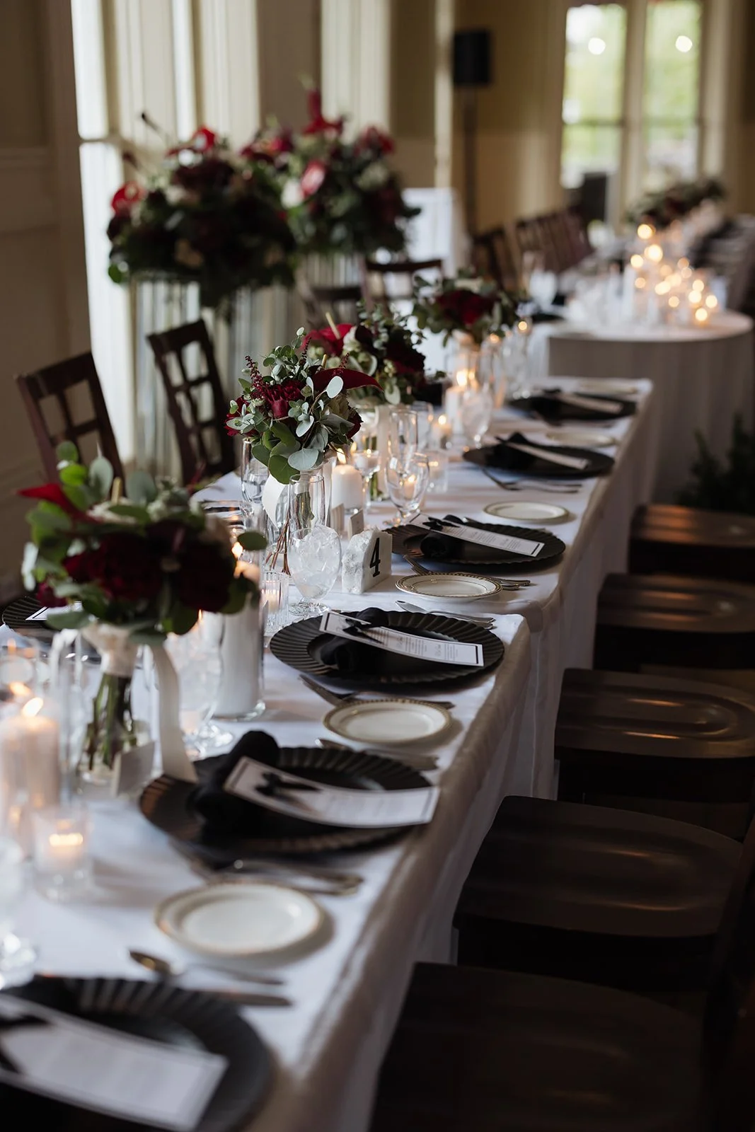 A long banquet table decorated with floral centerpieces, candles, plates, black napkins, and menus, set for a formal event in a well-lit room.