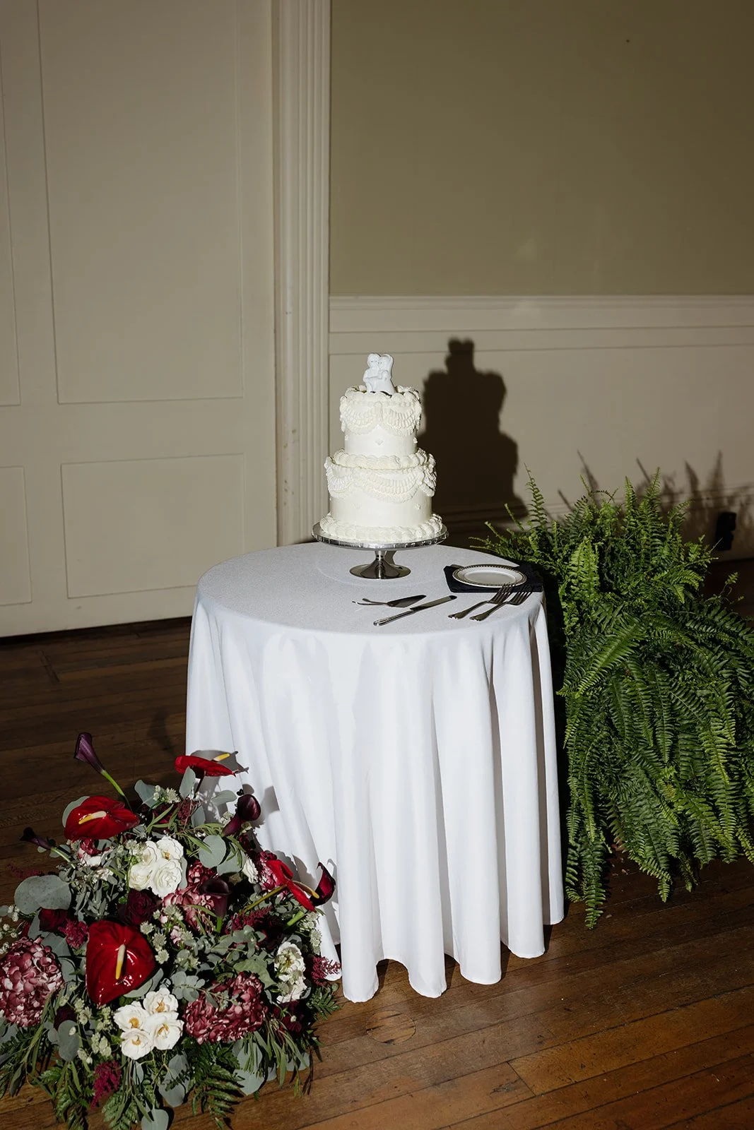 A white wedding cake with delicate lace-like decorations on top of a round table covered with a white tablecloth. Next to the cake is a small plate and utensils. There are colorful flowers and green foliage around the table.