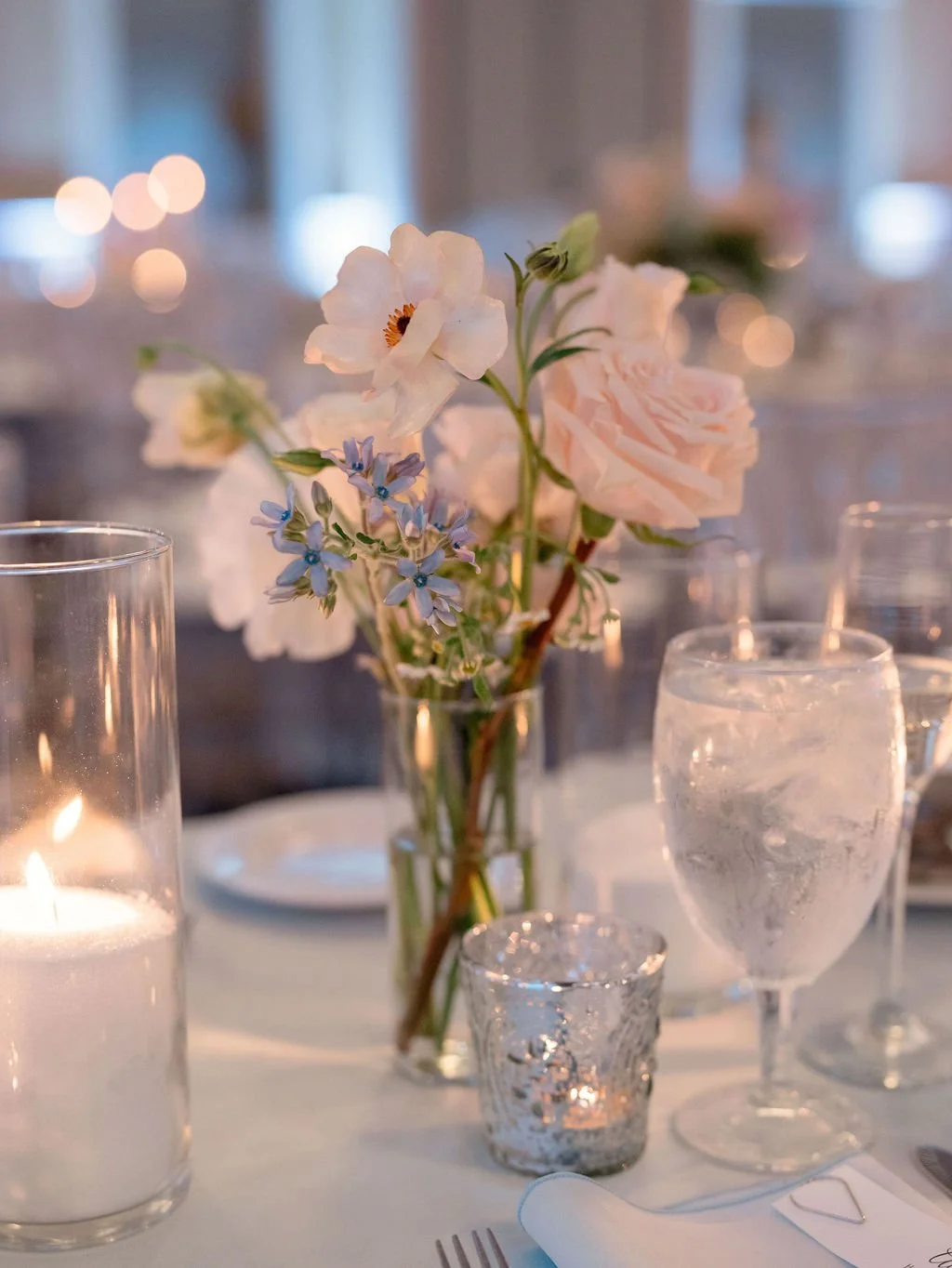 Elegant table centerpiece with soft pink roses, white and light purple flowers in a glass vase, surrounded by votive candles and glasses with water on a white tablecloth.