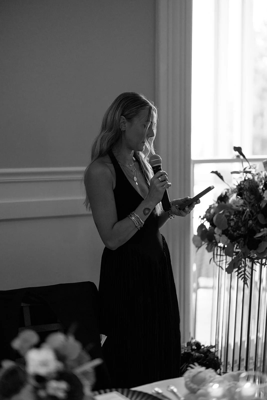 A woman with long hair, wearing necklaces and bracelets, standing indoors near a window, holding a microphone in one hand and a phone in the other, with flowers on a table nearby.