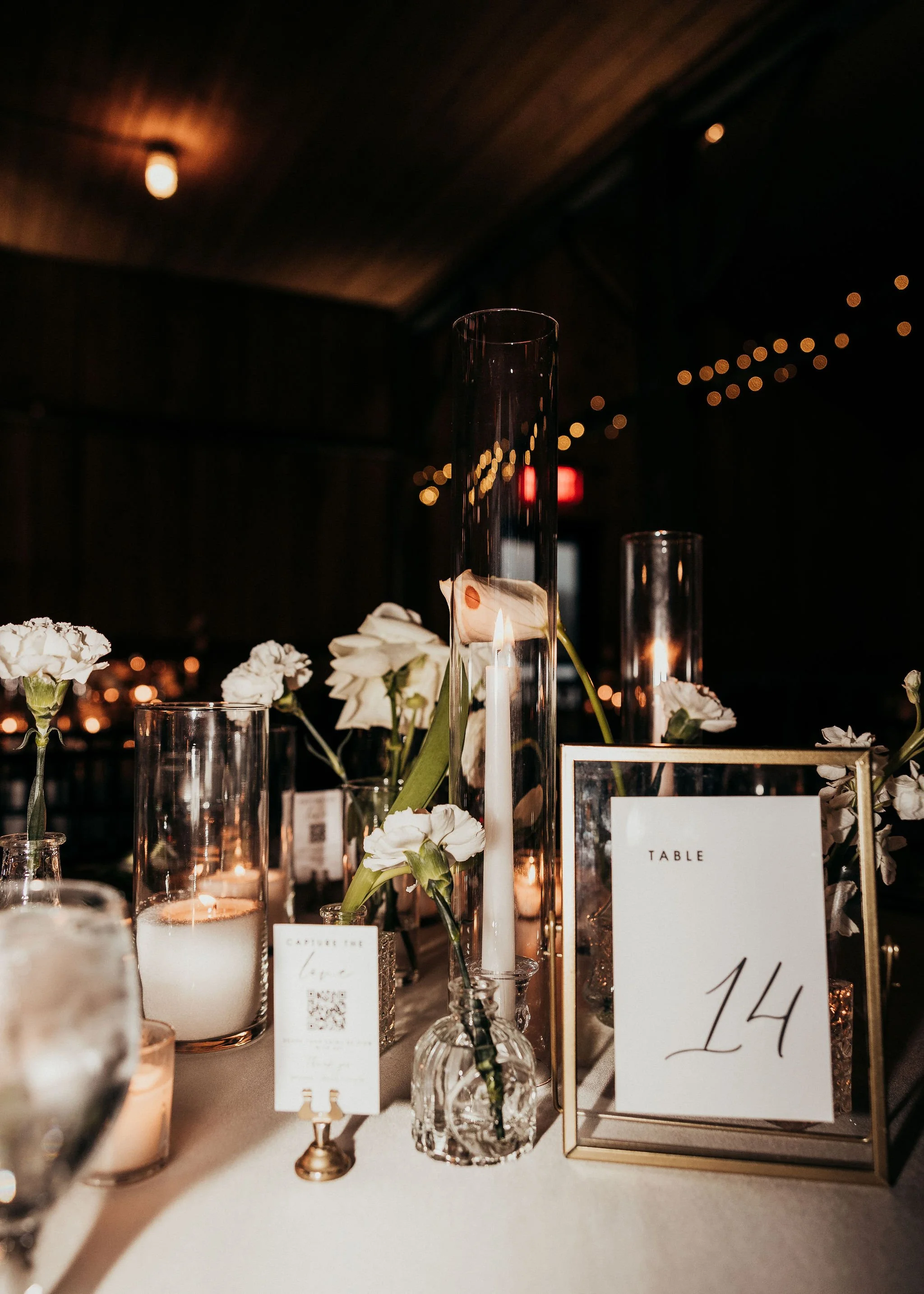 Elegant wedding table centerpiece with white flowers in glass vases, tall candle holders, and a table number card '14' on a beige tablecloth at a dimly lit venue with string lights.