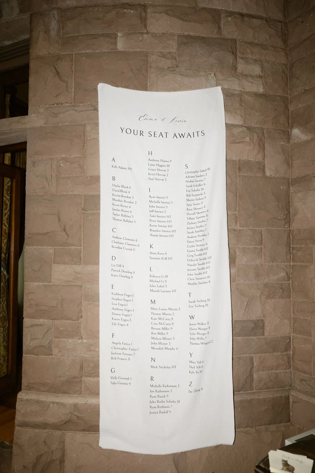 A large white poster with a list of guest names arranged alphabetically and assigned seat numbers, posted on a stone wall. The title reads: "Your Seat Awaits."