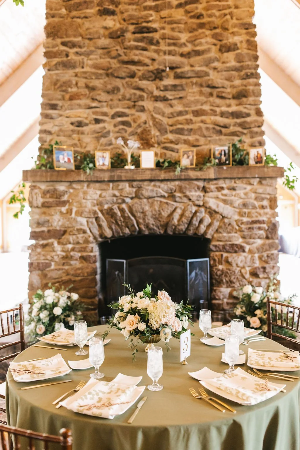 A decorated round table with a green tablecloth, set for six people with floral napkins, gold cutlery, and water glasses. The centerpiece is a large floral arrangement with white and blush flowers. Six chairs surround the table. Behind it, there is a