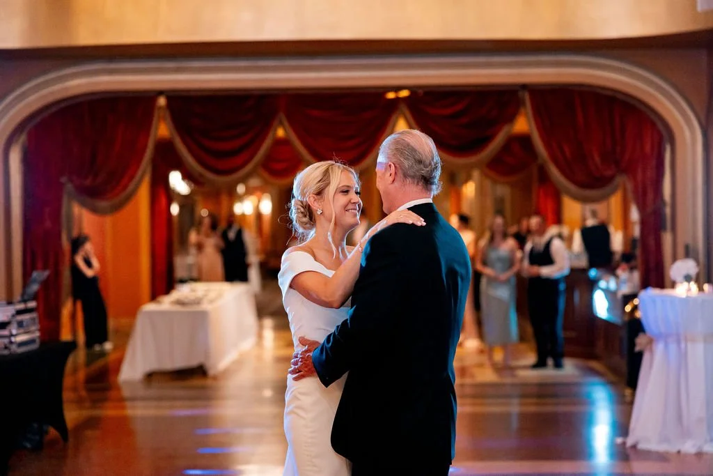 A bride and an older man dancing in a wedding reception hall, with red curtains and several guests in the background.