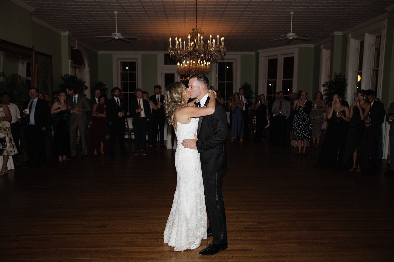A bride and groom sharing their first dance at a wedding reception, surrounded by seated and standing guests under large chandeliers in an elegant ballroom.