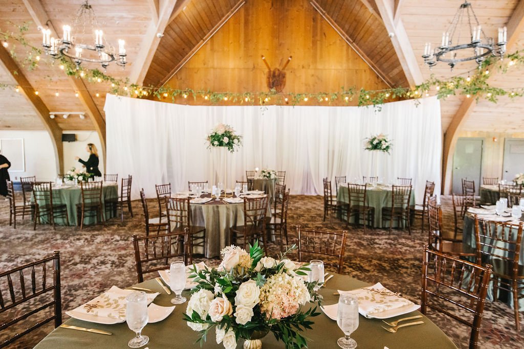 Wedding reception setup with round tables, green tablecloths, floral centerpieces, and white curtains background inside a wooden barn with chandeliers and greenery decor.
