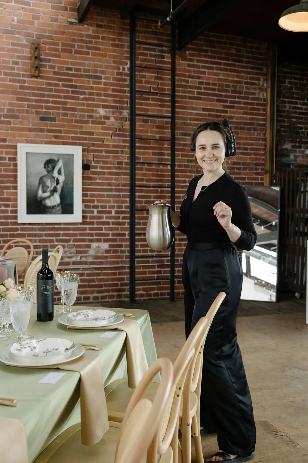 A woman in black attire with headphones, holding a teapot and smiling, standing in a restaurant with a brick wall background and elegantly set tables.