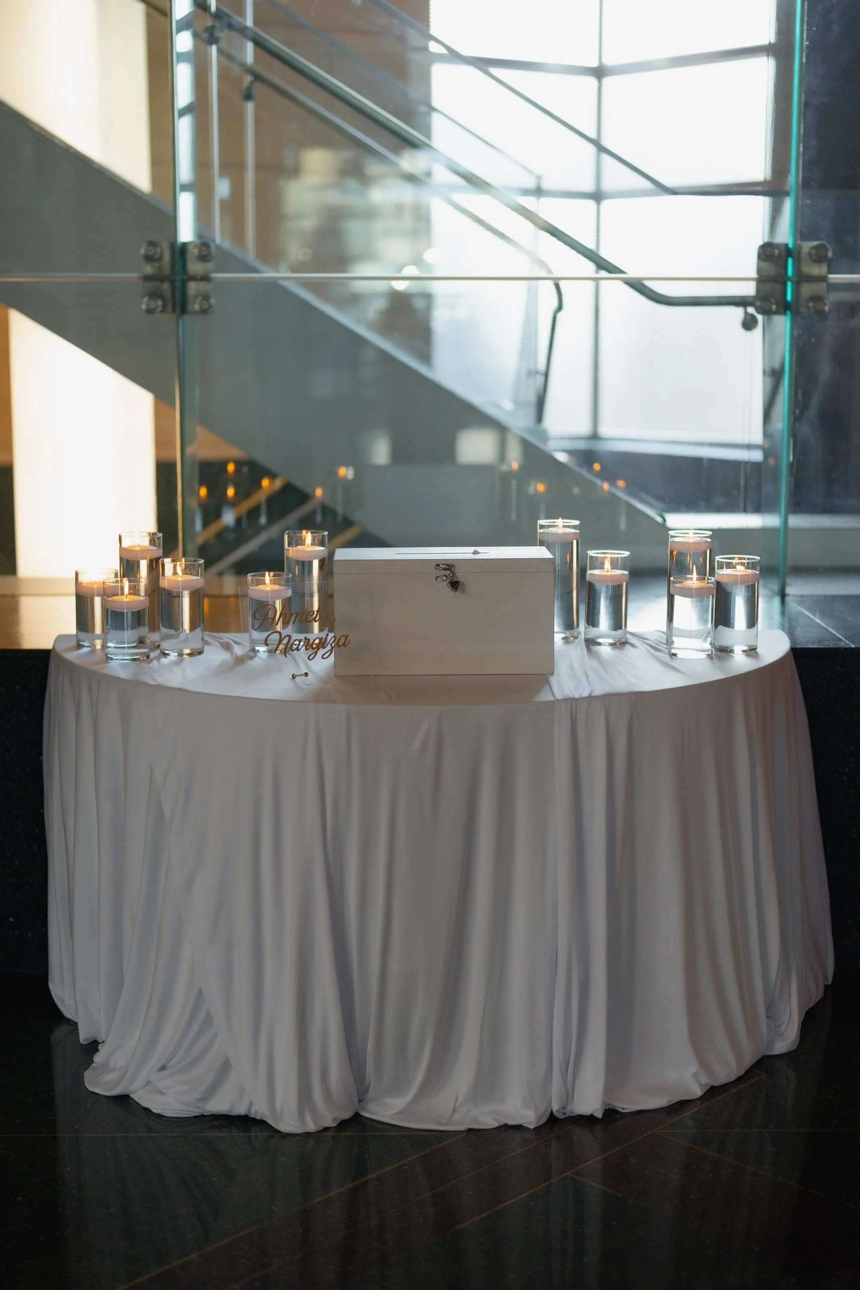 Table with a white tablecloth, candles in glass holders, a white wooden box with a lock, and a decorative sign with gold writing that says "Ahmet" and "Nargiza". The background shows a modern staircase with glass railings and metal supports.