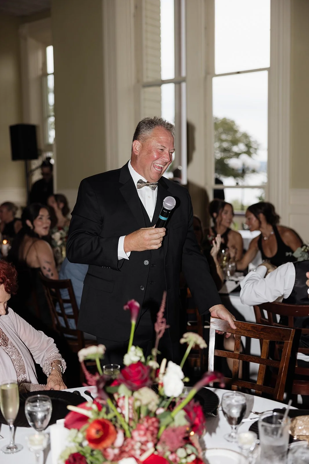 A man in a tuxedo with a bowtie and vest is smiling and speaking into a microphone at a formal event. The room has large windows, and there are guests seated at tables decorated with floral centerpieces.