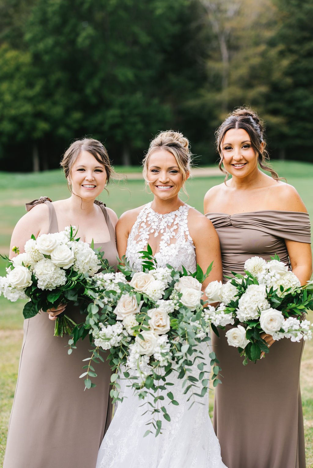 Three women, including a bride in a white lace wedding dress and two bridesmaids in taupe dresses, standing outdoors with a background of green trees and grass, holding large bouquets of white flowers and greenery.