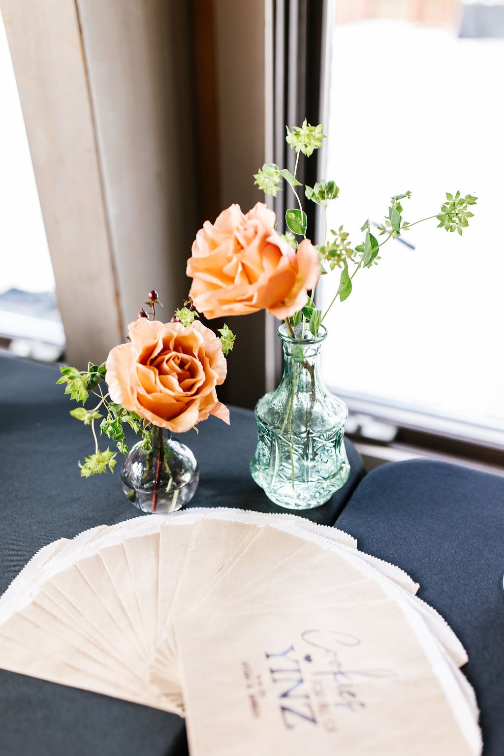 Two glass vases with peach-colored roses and green foliage on a black table near a window with white curtains, with folded paper fan in the foreground.