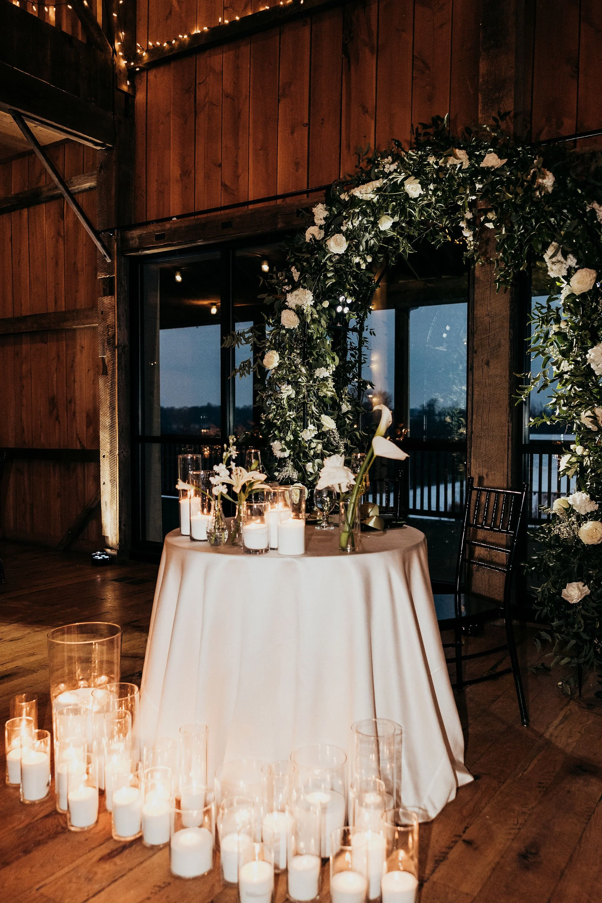 Indoor wedding or event space with wooden walls, string lights, a round table with a white tablecloth and candles, and a large floral arch.