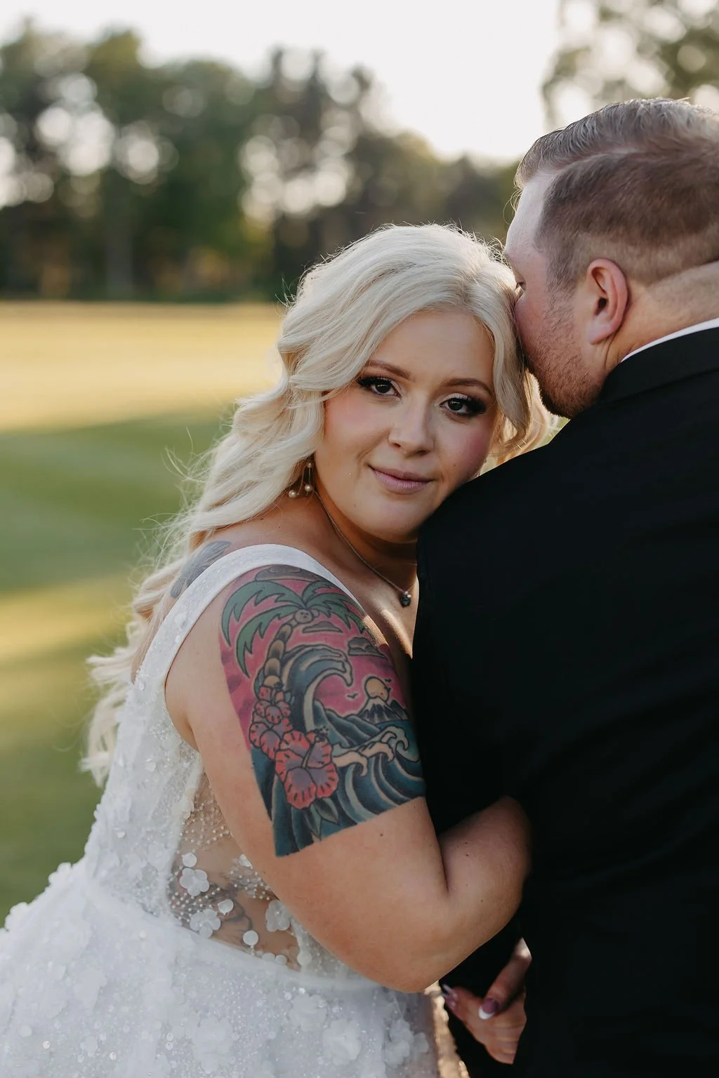 A bride with long blonde hair and a tattoo on her shoulder, wearing a white wedding dress, embraces a groom in a black tuxedo, with an outdoor background of trees and grass.
