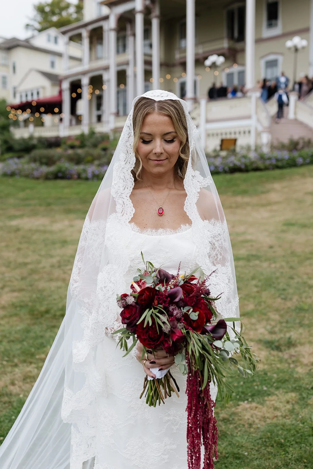 A bride in a white lace wedding dress holding a bouquet of dark red and purple flowers with greenery. She has a lace veil and wears a pendant necklace, standing outdoors in front of a house with a veranda decorated with string lights.