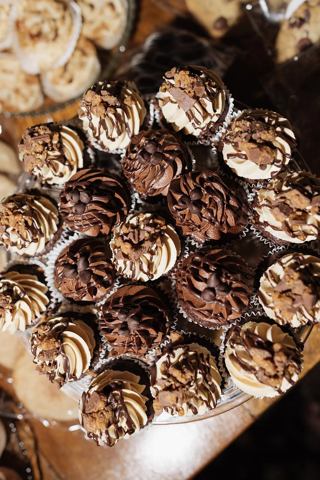 Close-up of a cake stand filled with assorted chocolate truffles, some topped with chopped nuts and drizzled with chocolate. In the background, there are some cookies or similar baked goods.