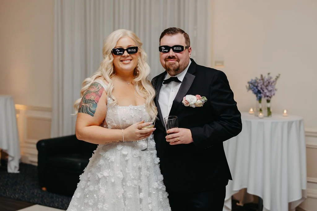 Bride and groom wearing sunglasses with text, smiling at their wedding reception, holding drinks, woman in a white wedding dress with tattoos, man in a black tuxedo, decorated reception area with flowers and candles.