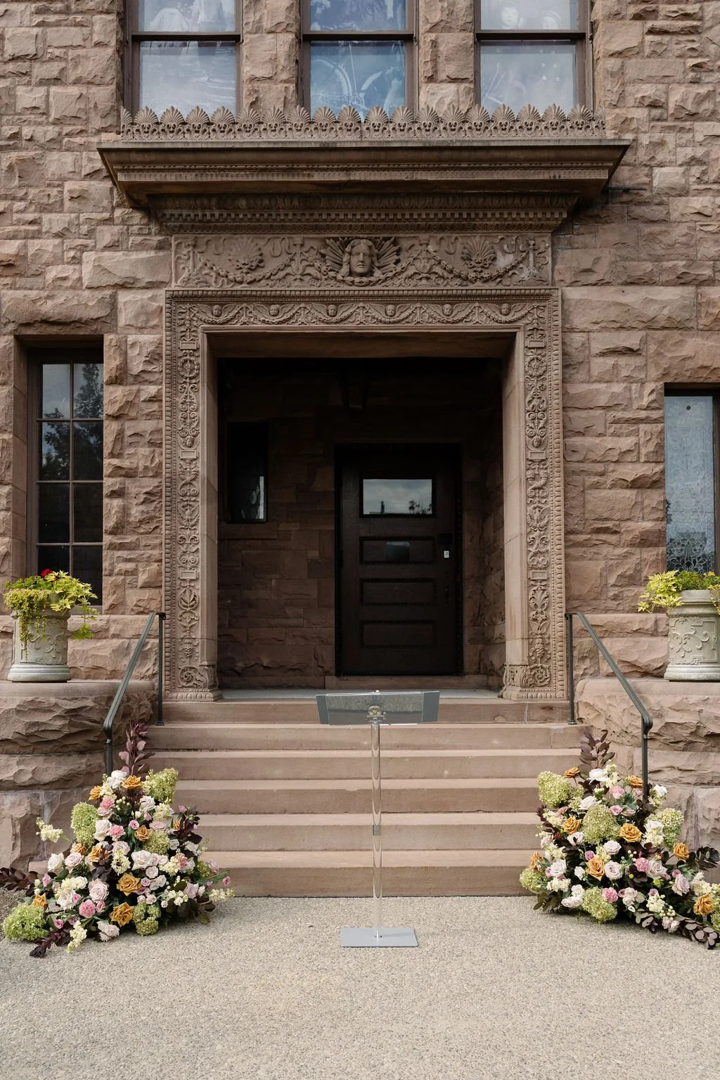 Stone building entrance decorated with floral arrangements on both sides of the steps, ornate stone carvings around the doorway, and a black door at the top of the stairs.