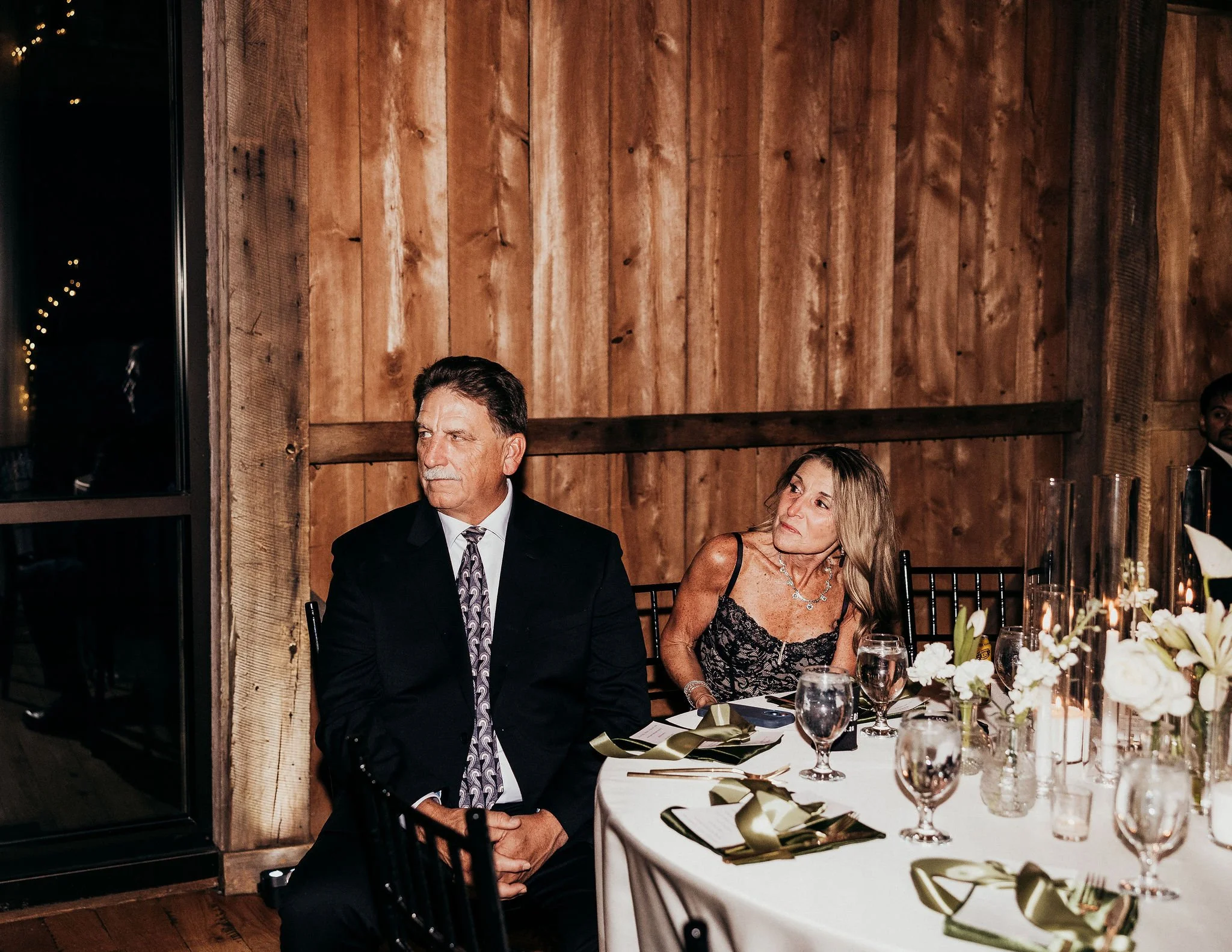 People sitting at a formal dinner event in a rustic wood-paneled room with elegant table settings and floral centerpieces.