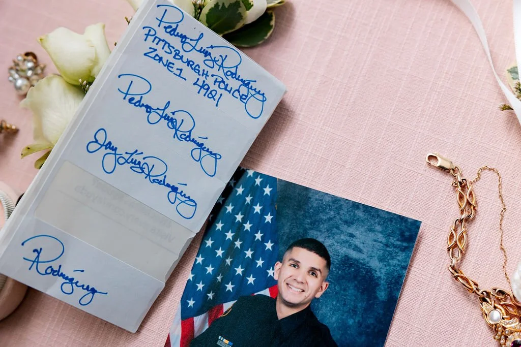 A graduation photo of a young man in a military uniform with an American flag in the background, along with jewelry and handwritten notes on a pink tablecloth, surrounded by white roses and decorative items.