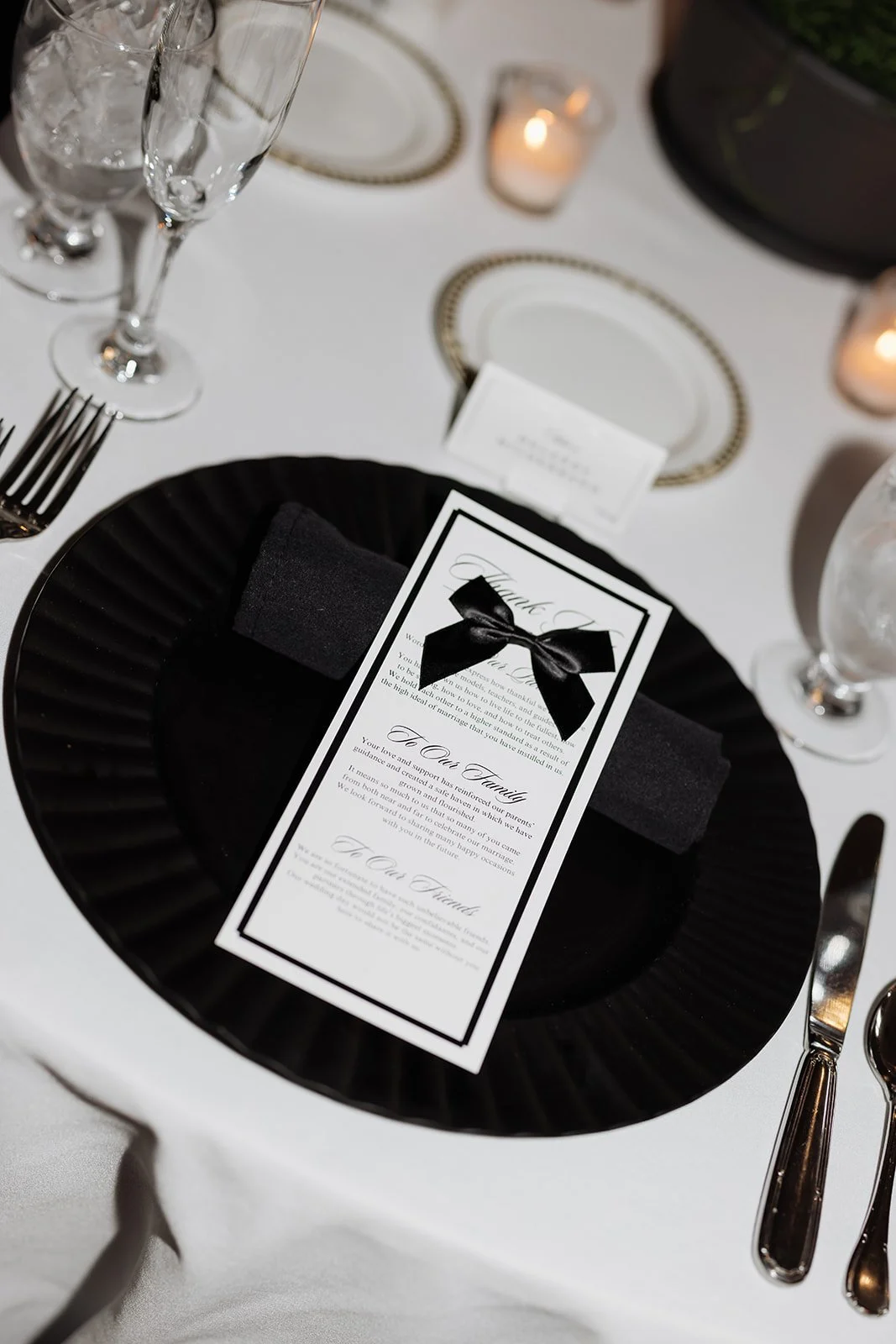 Elegant table setting featuring a black plate with a black napkin wrapped around a wedding menu card decorated with a black bow, alongside silverware, wine glasses, a lit candle, and a printed name card.