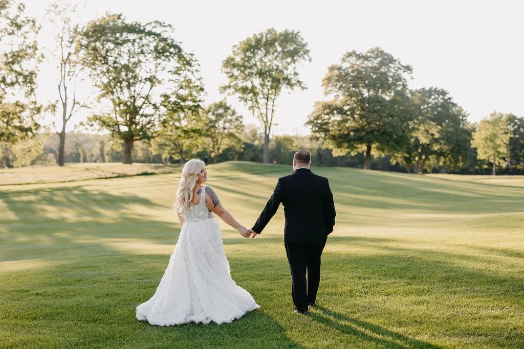 A bride and groom holding hands and walking away on a lush green golf course during sunset, surrounded by trees.