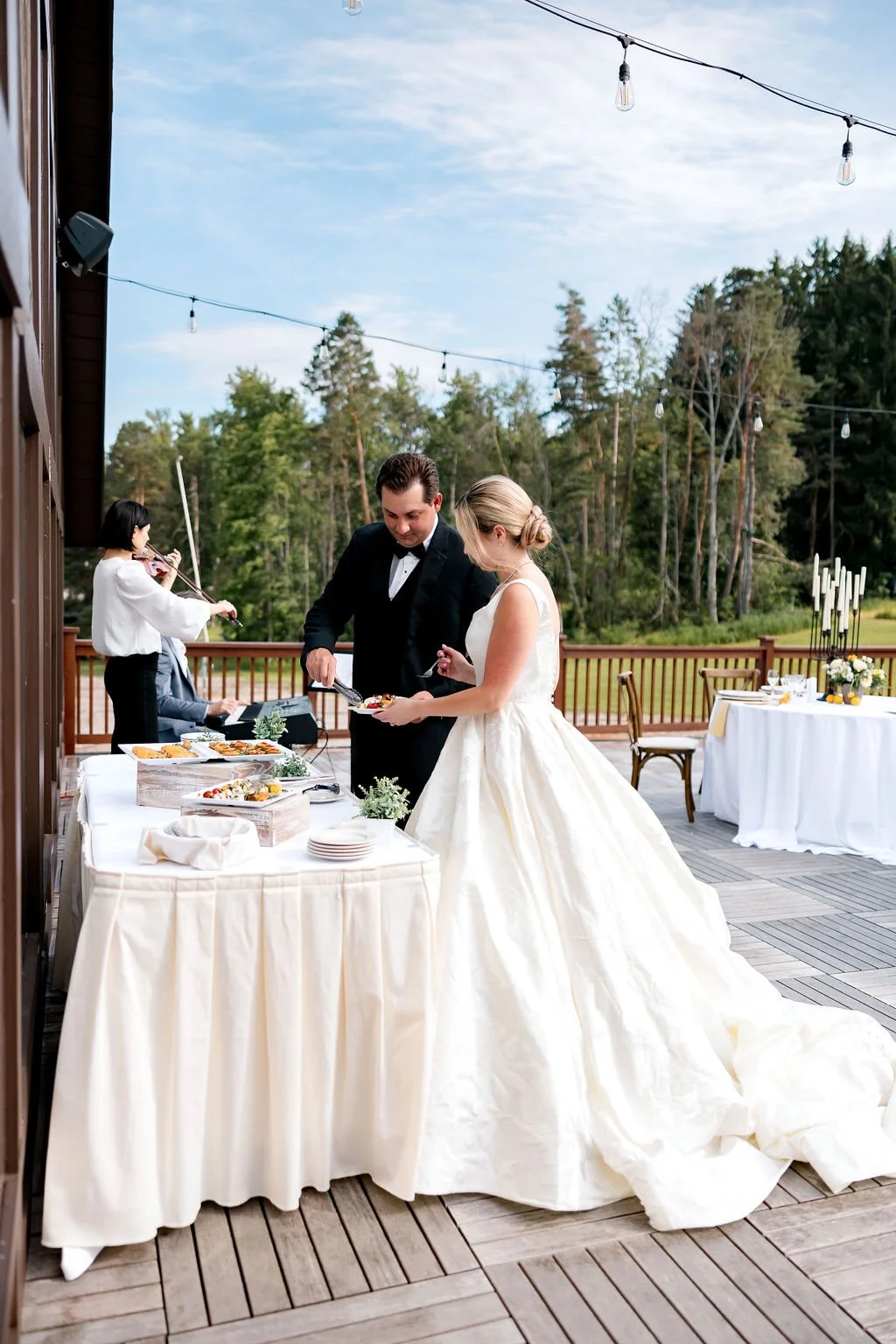 Bride and groom at an outdoor wedding reception, serving themselves from a buffet table with a string of hanging lights overhead and trees in the background.