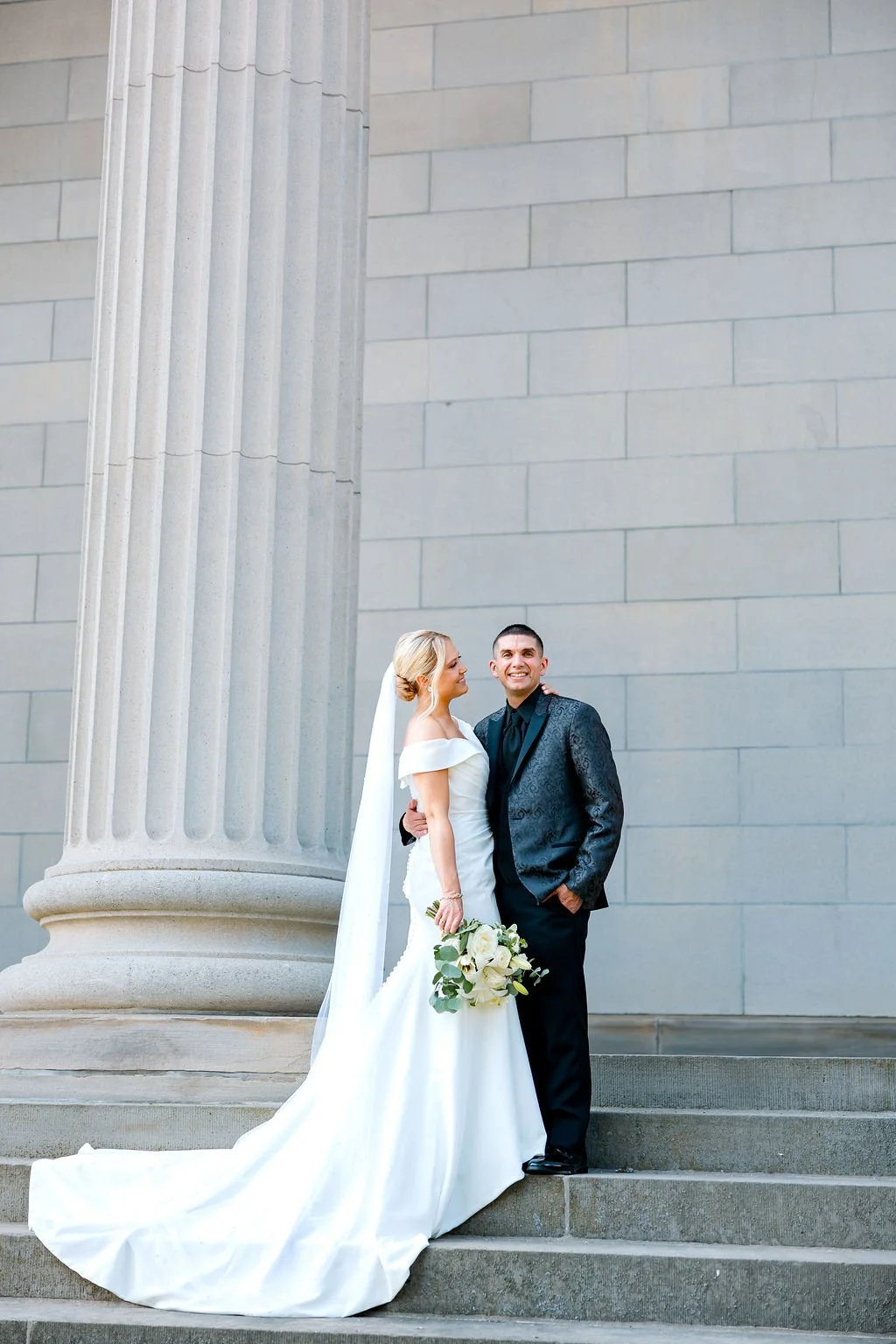 Bride and groom standing together on steps outside a building with large columns, smiling, with the bride holding a bouquet of white flowers.
