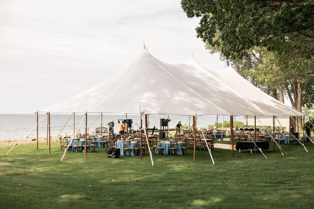 Large white event tent over a grassy outdoor area near a body of water, with tables and chairs set up underneath and people preparing musical equipment