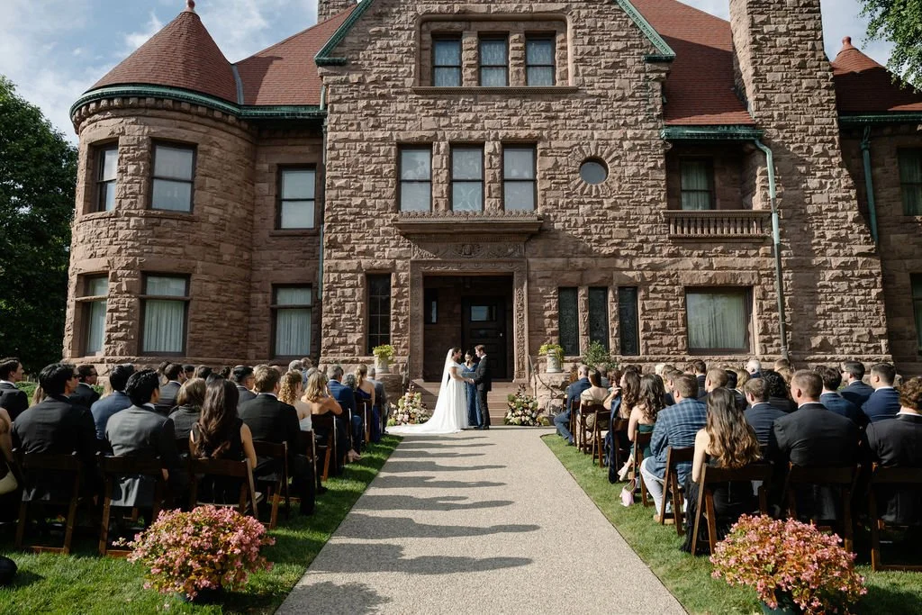 A wedding ceremony taking place outside a large, brown stone mansion with a red-tiled roof. The bride and groom are standing in front of the mansion, facing each other. Guests are seated on either side of the aisle, watching the ceremony. There are p