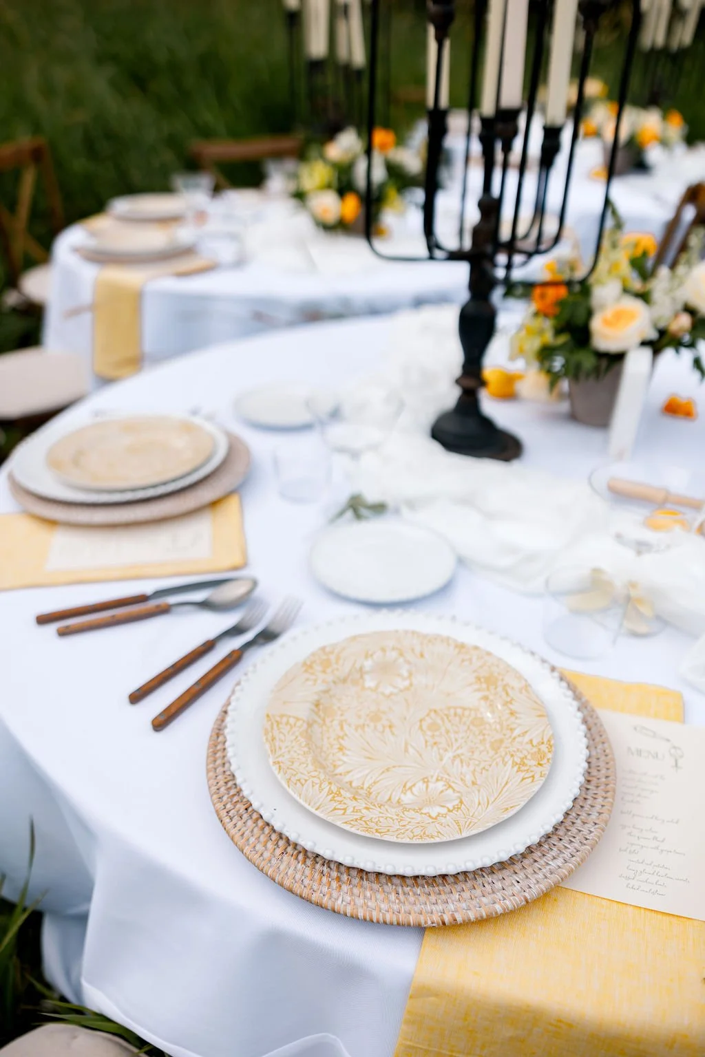 Elegant outdoor dining table set with white tablecloth, yellow accents, decorative plates, cutlery, glasses, and floral centerpiece, surrounded by chairs and greenery.
