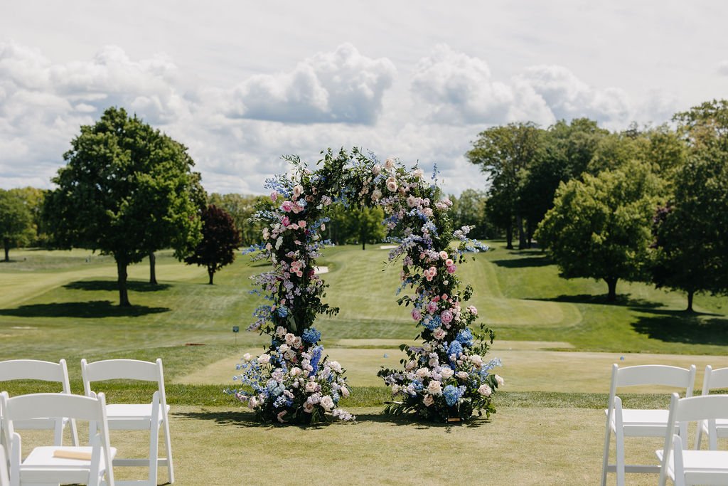 Wedding ceremony setup with a floral arch and white chairs on a grassy field with trees and cloudy sky in the background.
