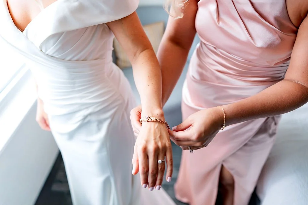 A bride in a white wedding dress having jewelry fastened by another woman in a pale pink dress.