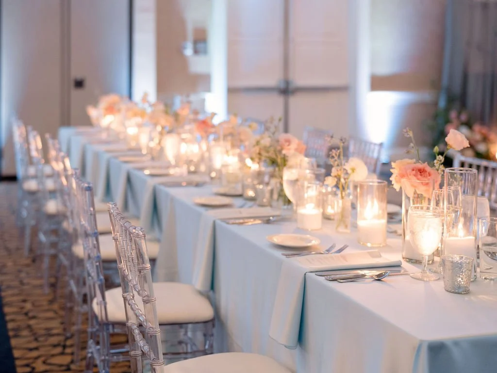Elegant long dining table with pastel pink and white floral centerpieces, candles, and place settings with white plates, silverware, and glassware in a softly lit room, ready for a formal event.