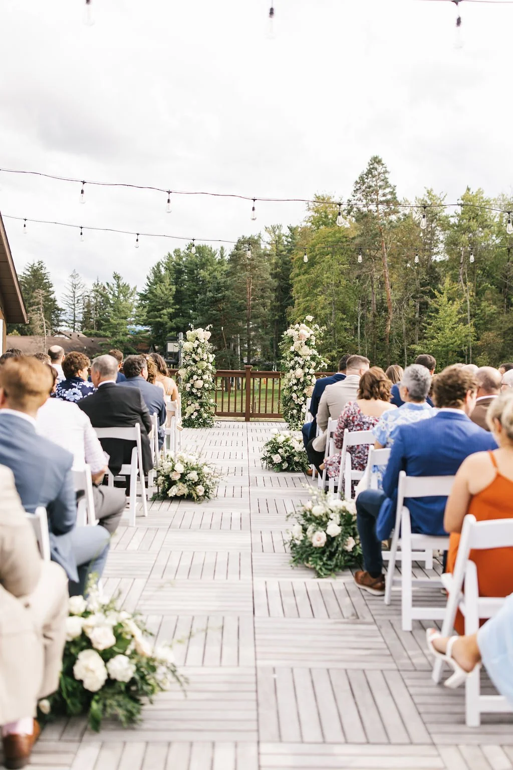 Outdoor wedding ceremony with rows of seated guests facing a decorated altar with white flowers, on a wooden deck surrounded by trees and string lights overhead.