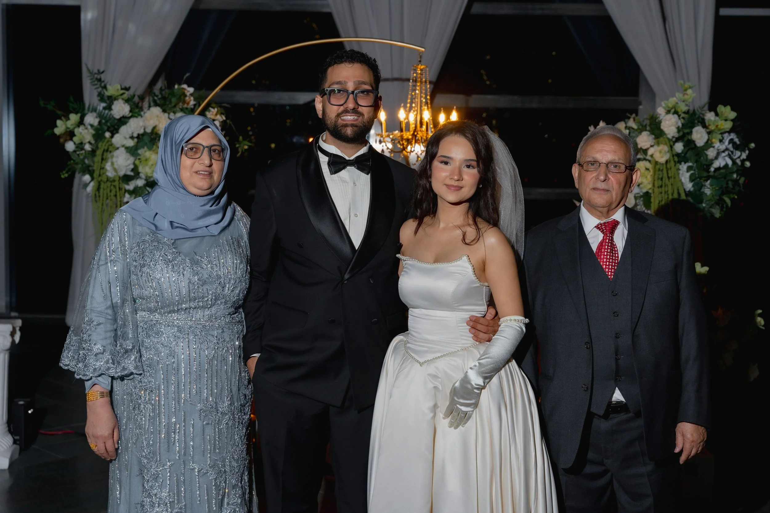 A group of five people at a wedding reception, including a woman in a light blue dress and hijab, a man in a black tuxedo, a woman in a white wedding gown, an older man in a dark suit, and an older man in a dark suit with a red tie, standing together