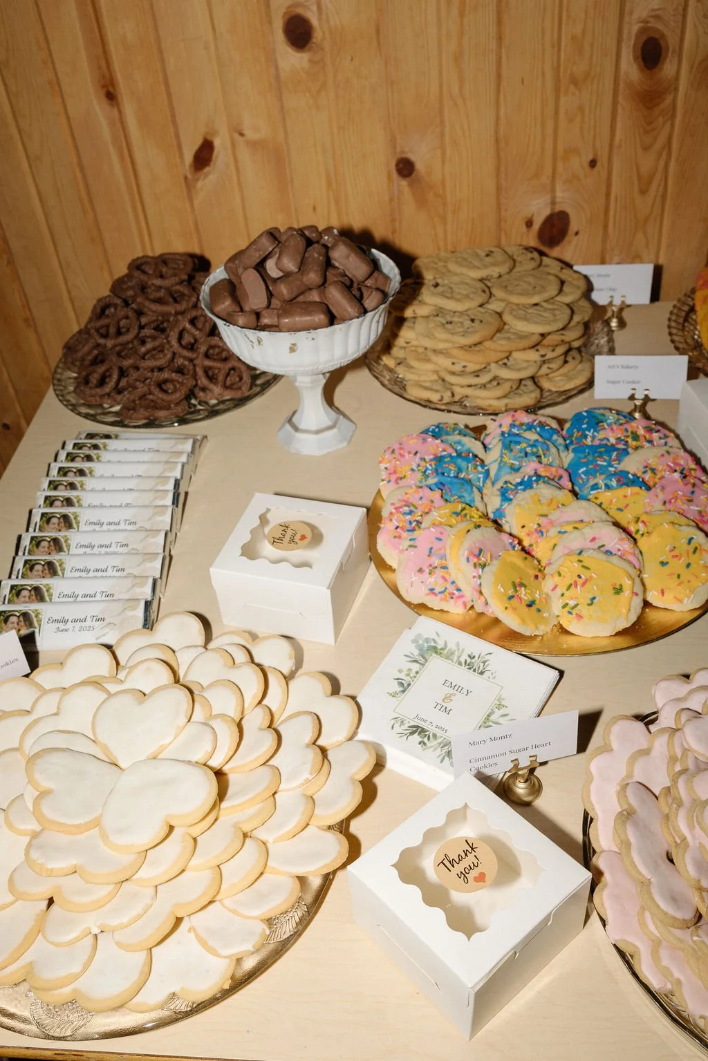 Display of assorted cookies, chocolates, and candy on a table at an event, with small signs and personalized wedding favors.